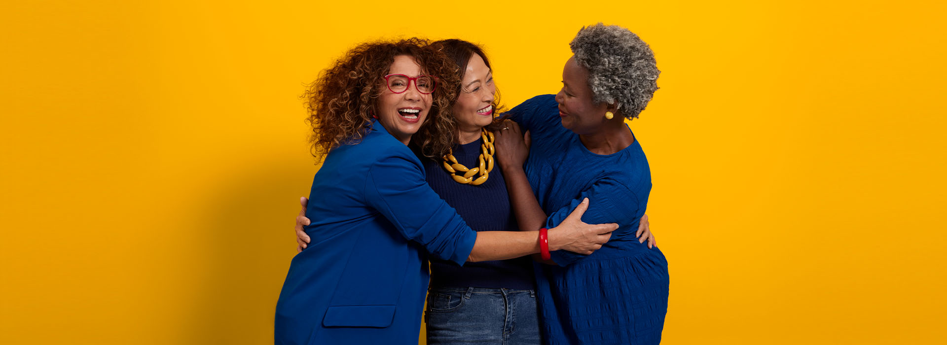 Three middle-aged women of different ethnicities dressed in blue hugging and smiling.