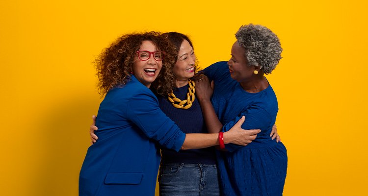 Three middle-aged women of different ethnicities dressed in blue hugging and smiling.  